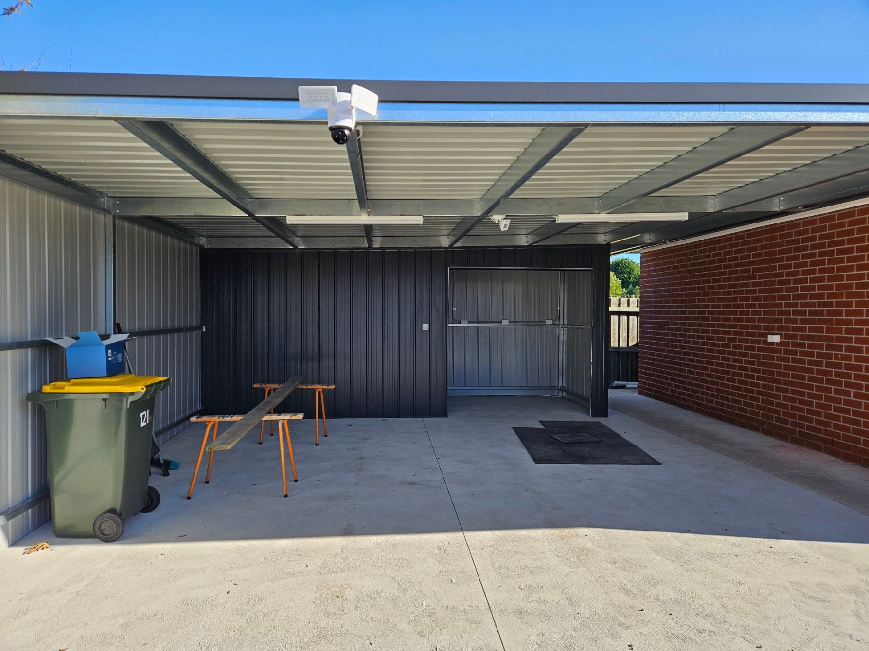 A carport with a green trash can and a brick wall