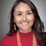 Woman smiling, wearing a red top and beaded necklace, dark hair against a gray background.