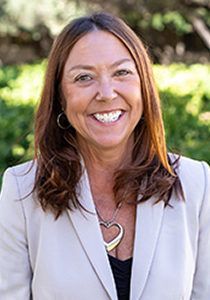 Woman with brown hair smiles in a beige blazer, a heart pendant, with greenery blurred in the background.