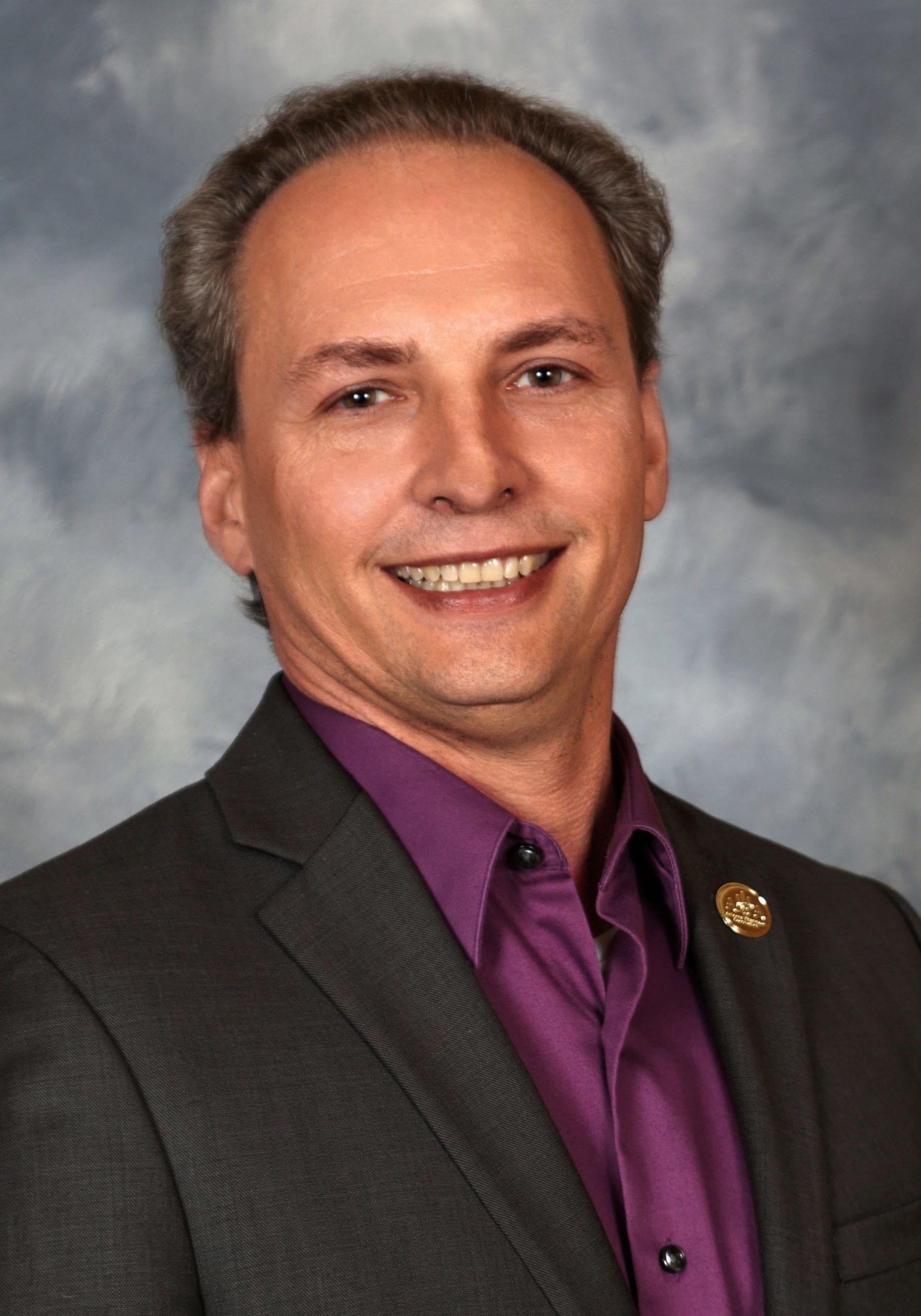 Man in suit jacket and purple shirt smiles; background is blue and gray.