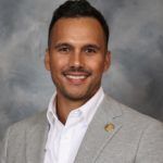 Man in gray blazer and white shirt smiling. Background is gray and blue.