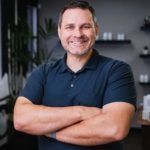 Man with arms crossed smiling in a modern office. He is wearing a dark blue polo.