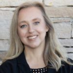 Blond woman smiling, wearing a black blazer, standing in front of a stone wall.