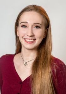 Woman with long blonde hair, smiling, wearing a red sweater and necklace.