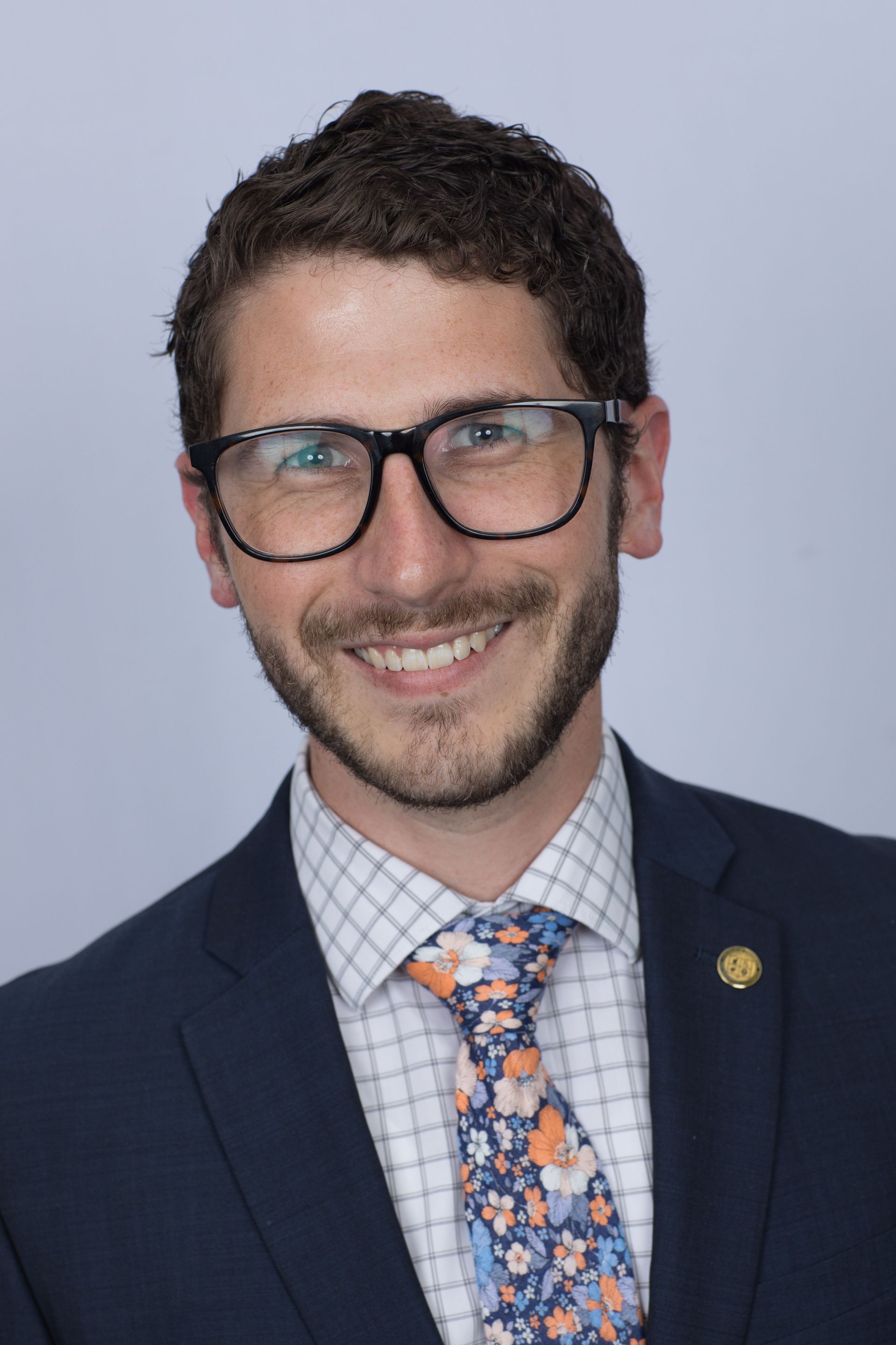 Man with glasses and a patterned tie smiles, wearing a suit jacket and collared shirt.