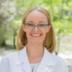 Woman in a white coat and glasses smiling in front of blurred greenery.