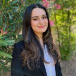 Woman in black blazer smiles outdoors, long brown hair, white shirt.