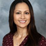 Woman with long brown hair, smiling, wearing a dark red polka dot shirt, against a blurred background.