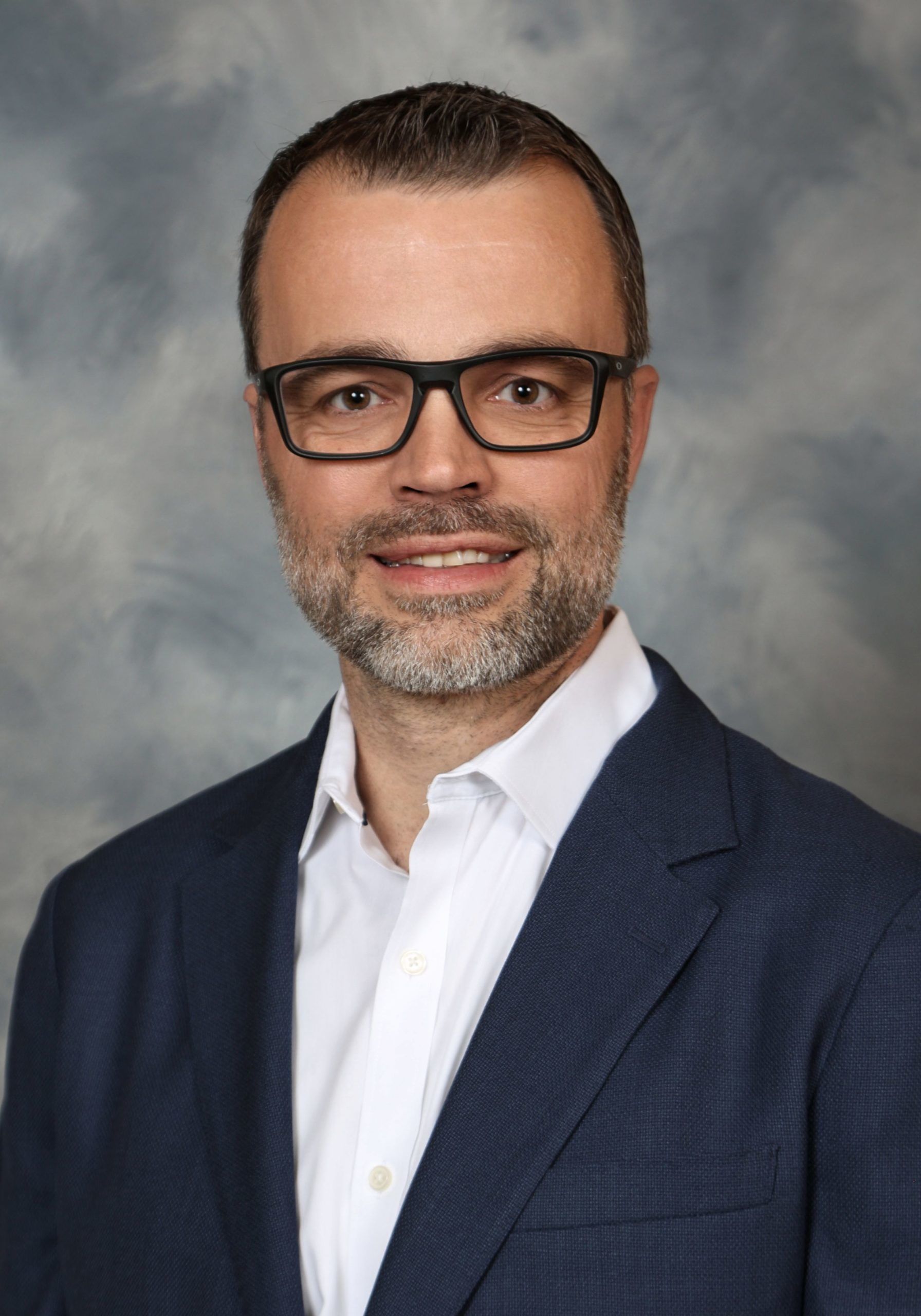 Man with glasses, wearing a blue blazer and white shirt, smiles at the camera. Neutral background.