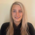 Woman with blonde hair smiling, wearing a black top against a light wall.