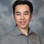Man in gray shirt smiles, posing against a blurry background.