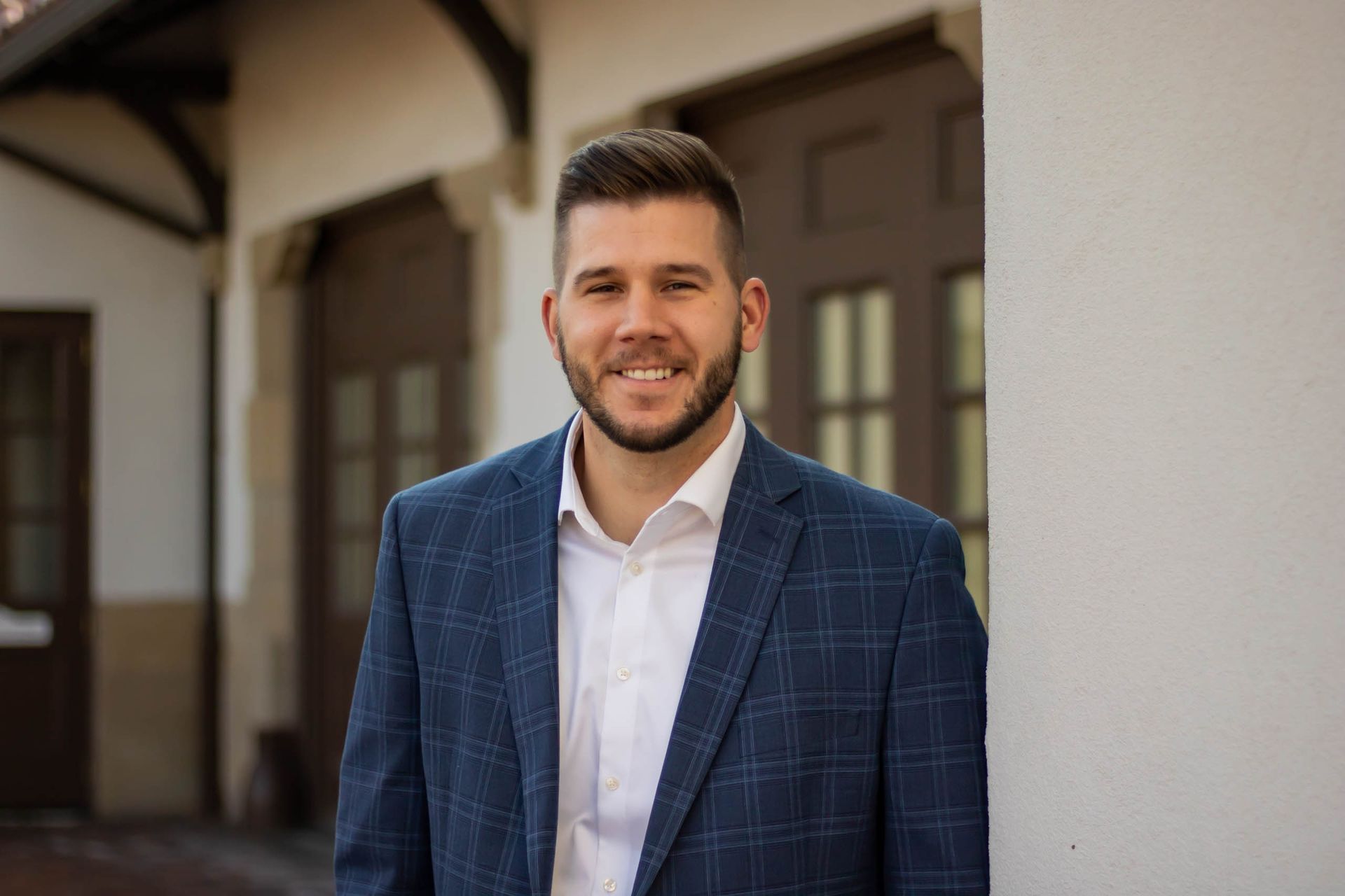 Man in blue blazer smiles, leaning against a white wall; background shows arched doorway with wooden doors.