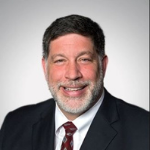 Man with a graying beard smiles, wearing a dark suit and a maroon tie.