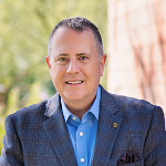 Man in a blue shirt and blazer, smiling outdoors with a blurred background.