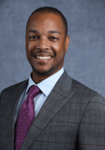 Man in a gray suit with a purple tie smiles in a professional headshot.