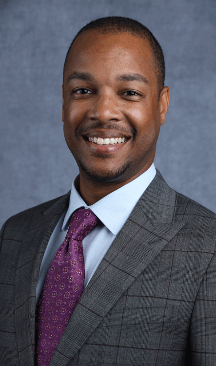 Man in a gray suit and purple tie smiling, light blue shirt, against a dark blue background.
