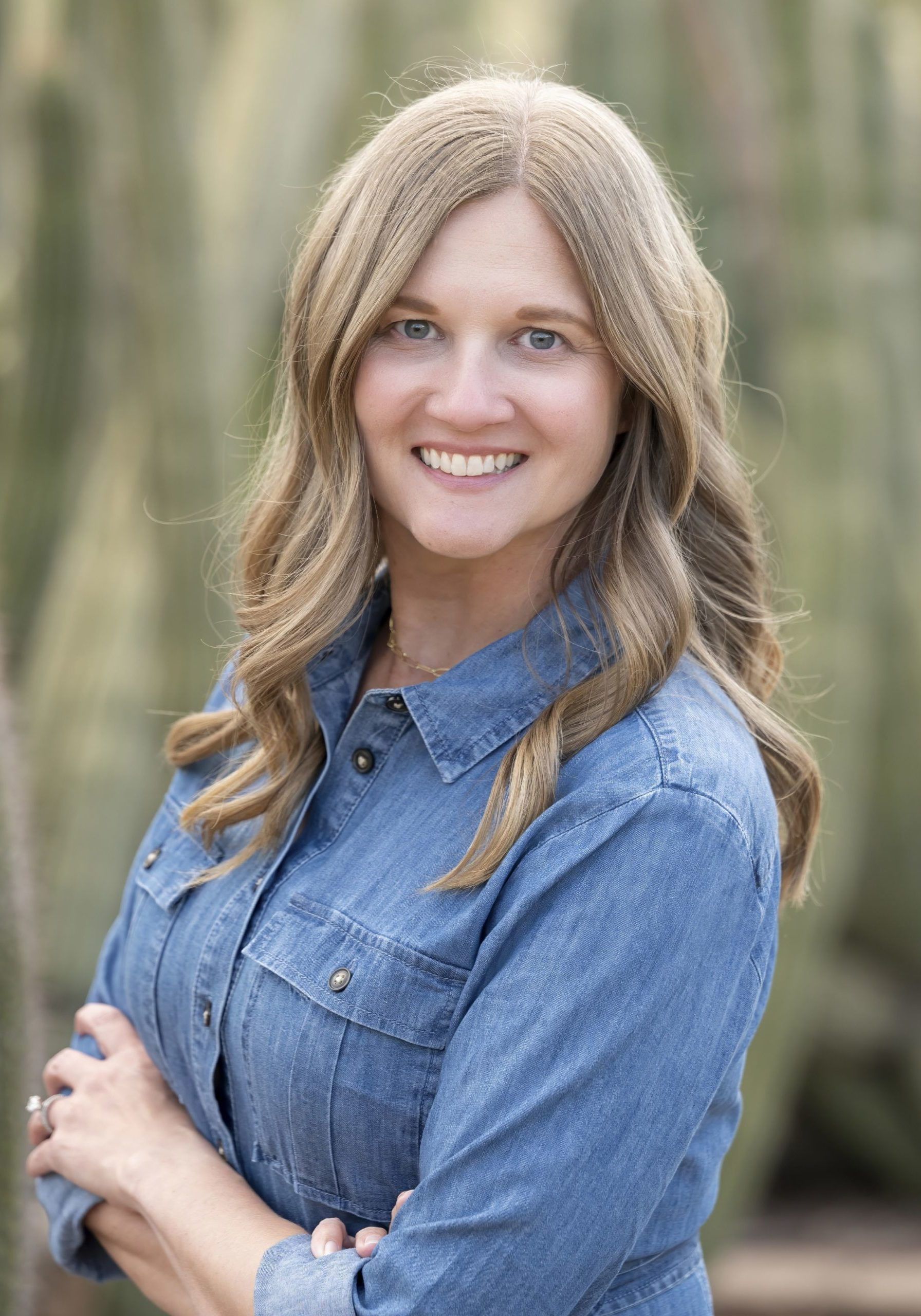 Woman smiling, arms crossed, wearing denim shirt, outdoors, blurred cactus background.