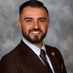 Man in brown suit smiles at the camera, light blue shirt, dark tie, formal portrait.