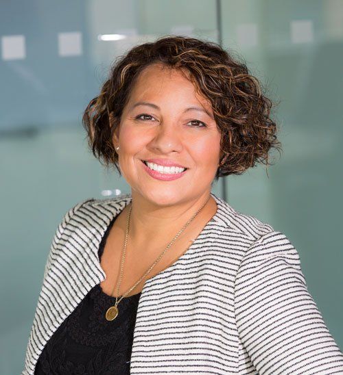 Woman smiling, wearing a black top and striped jacket, in a modern office.