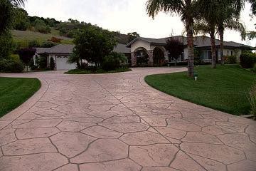 Beige stone-patterned driveway leading to a large house with a grassy lawn, trees, and a hillside background.