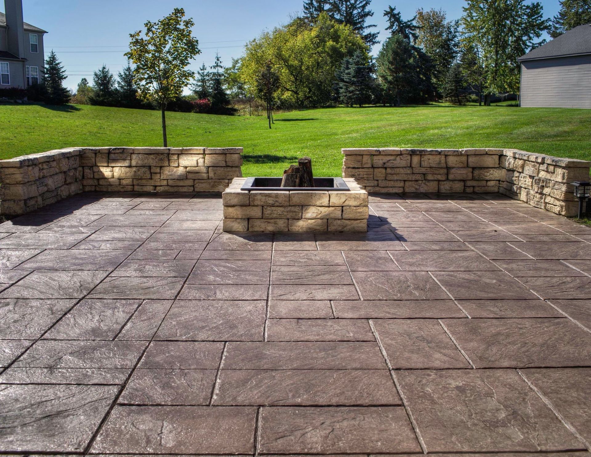 Stone patio with fire pit and low walls, surrounded by green grass and trees.