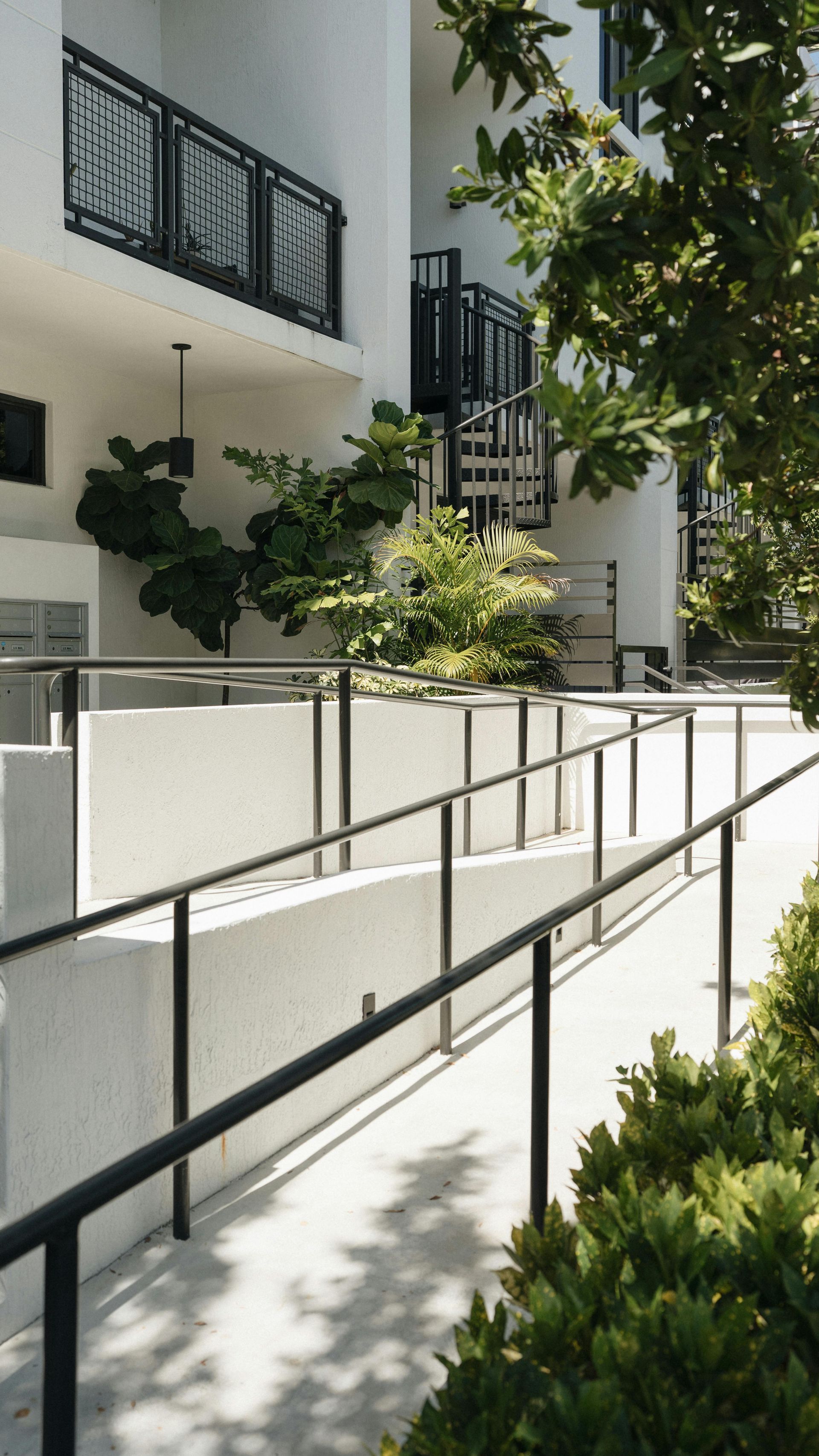 Exterior shot: ramp with black railing leads up to a white building with black balconies and a spiral staircase, surrounded by green foliage.