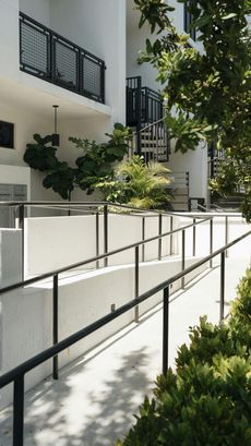 Exterior shot: ramp with black railing leads up to a white building with black balconies and a spiral staircase, surrounded by green foliage.