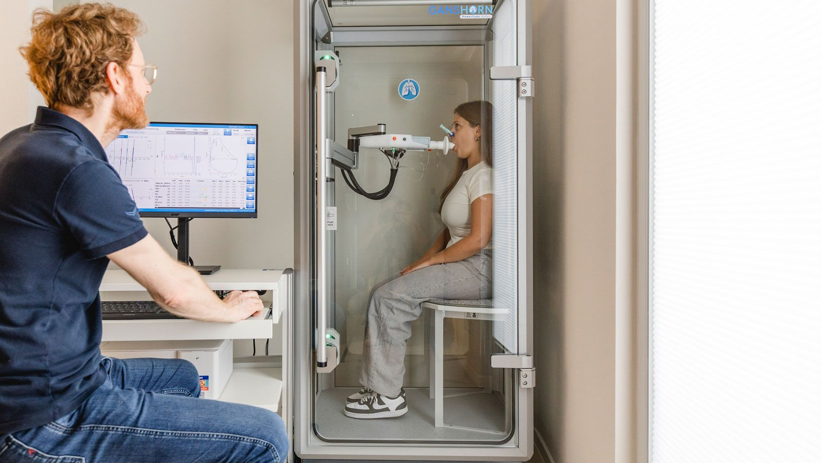 Bodyplethysmographie, Arzt sitzt am Coputer und überwacht die Ergebnisse, Frau sitzt in einer Kabine mit einem Mundstück