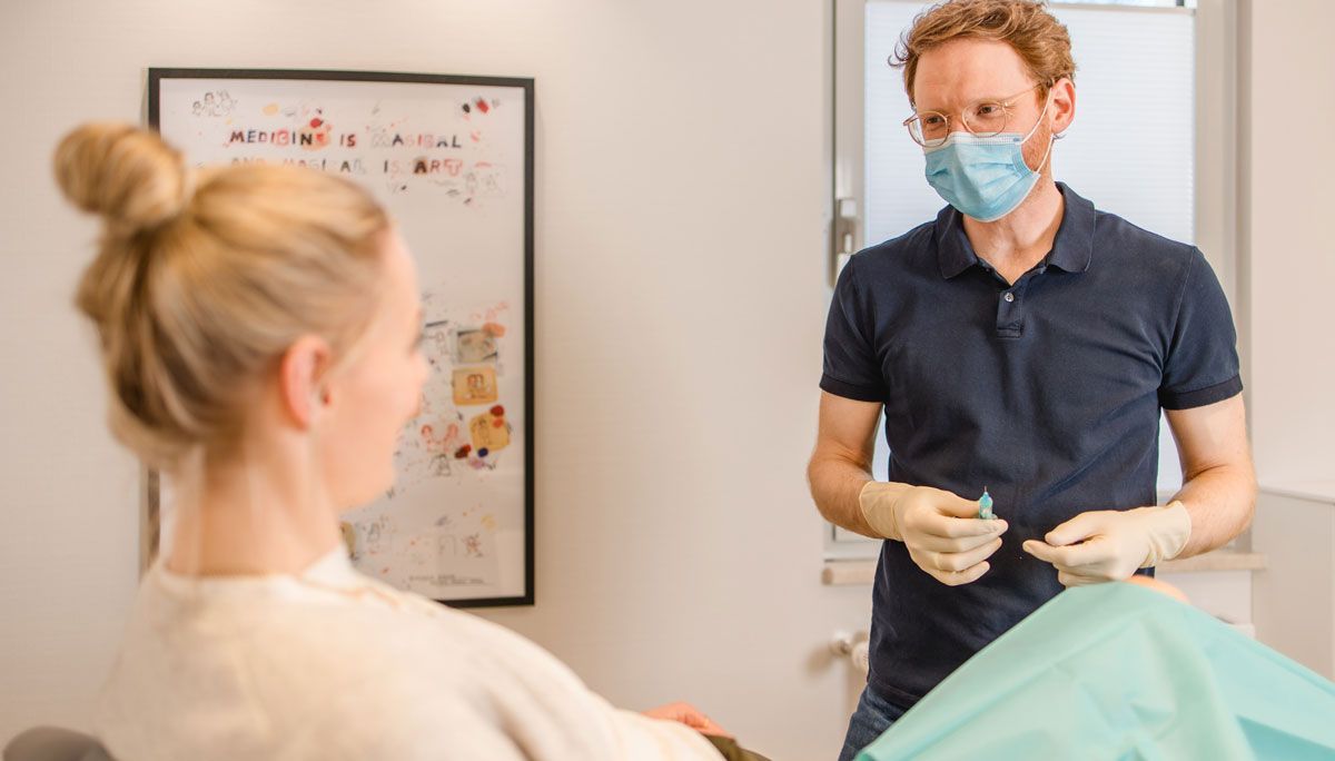 Praxis Kramer + Arndt, Dr. Kramer hält eine Spritze in der Hand, Patientin mit hochgezogenen Knie liegt auf der Wiege