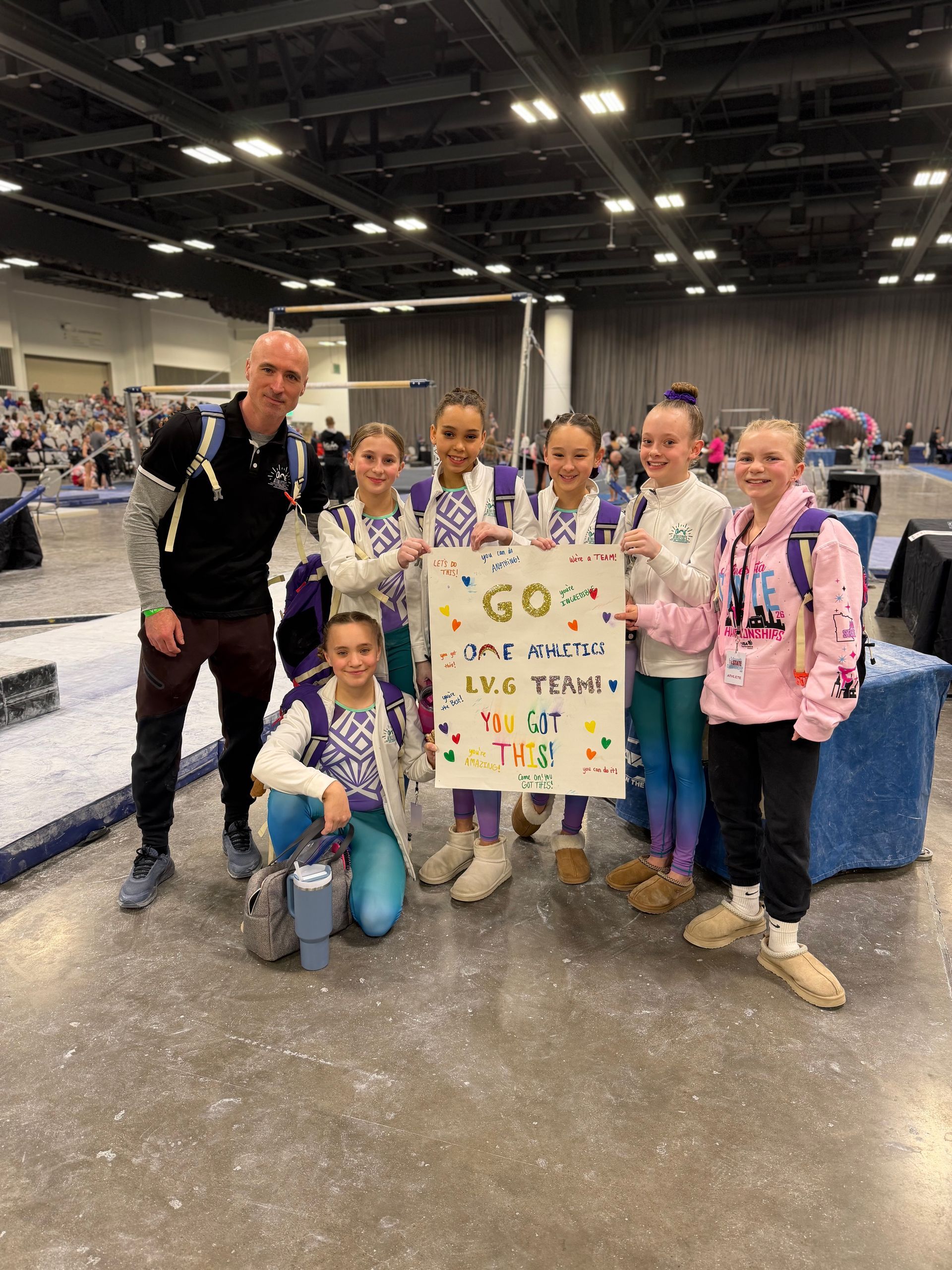 Group posing with a colorful sign at a convention center, standing and kneeling near tables under bright lights.