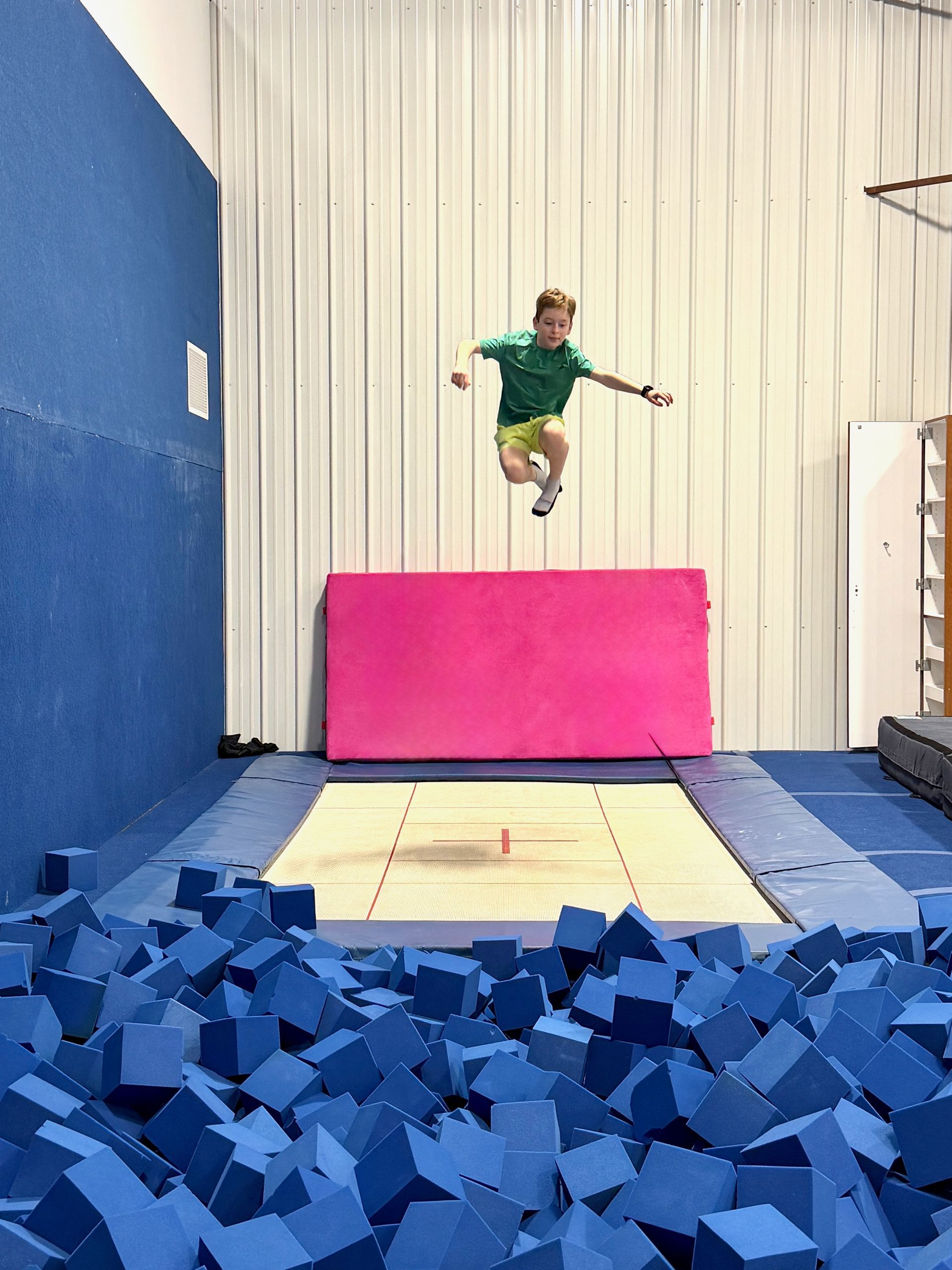 Child seated on a black gym machine in a weight room with speckled flooring and exercise equipment in the background