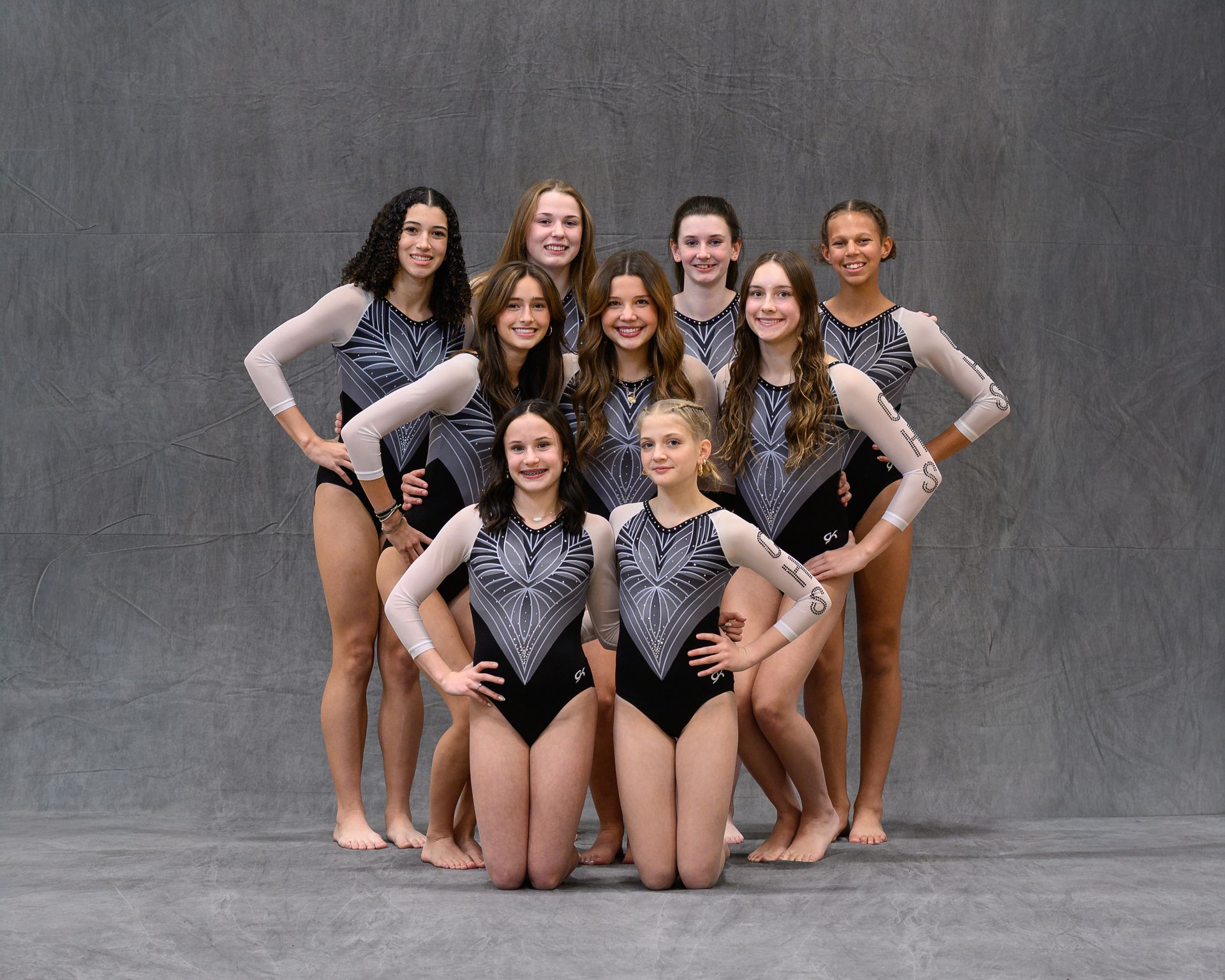 Gymnasts sitting in a line on a blue mat, watching a coach in an indoor gym.