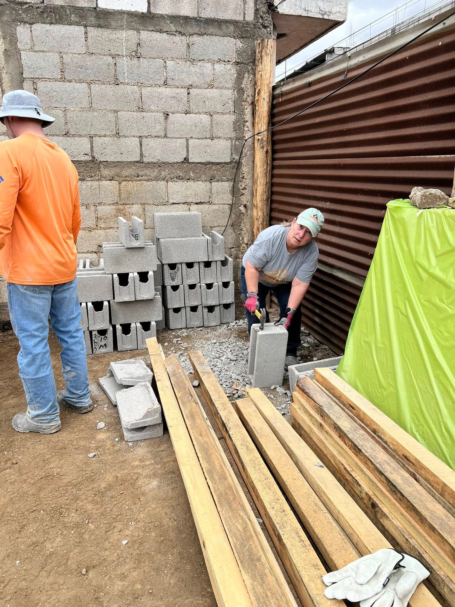 Two people working on a construction project with concrete blocks, wooden beams, and a corrugated metal wall.