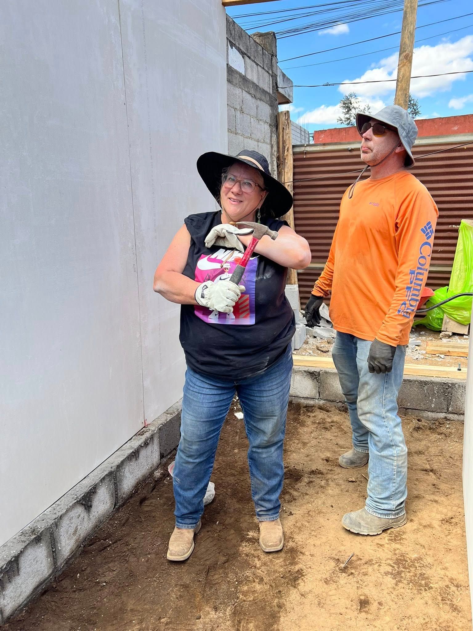 Two people in construction gear, one holding a hammer, stand near a partially finished white wall at a building site.