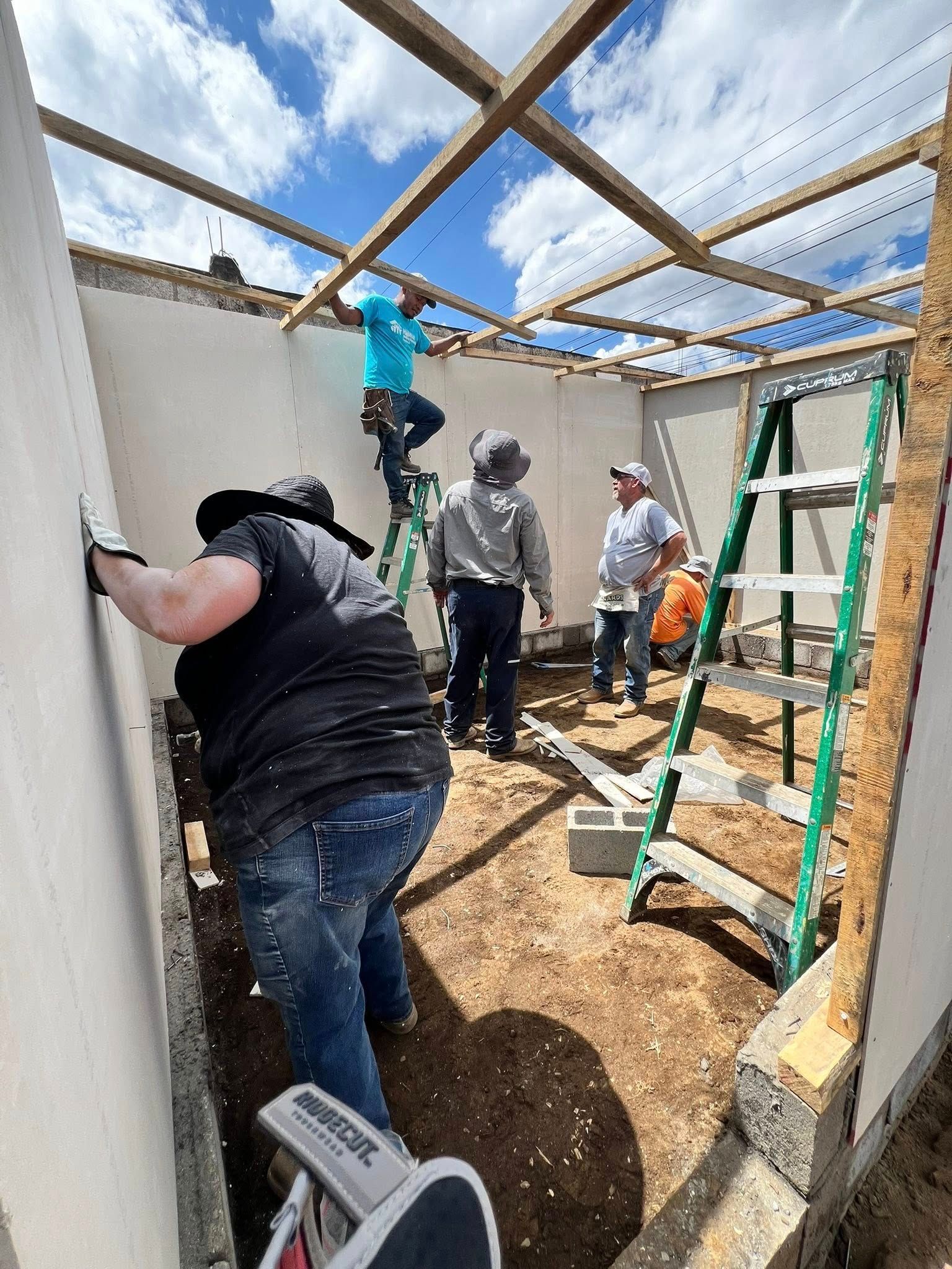 Four people working together to build the wooden frame and walls of a structure on a dirt-floored site.