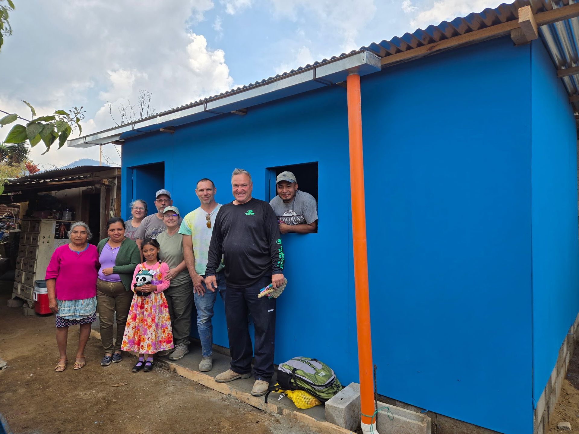 A group of people stands in front of a newly painted bright blue house with a corrugated metal roof.