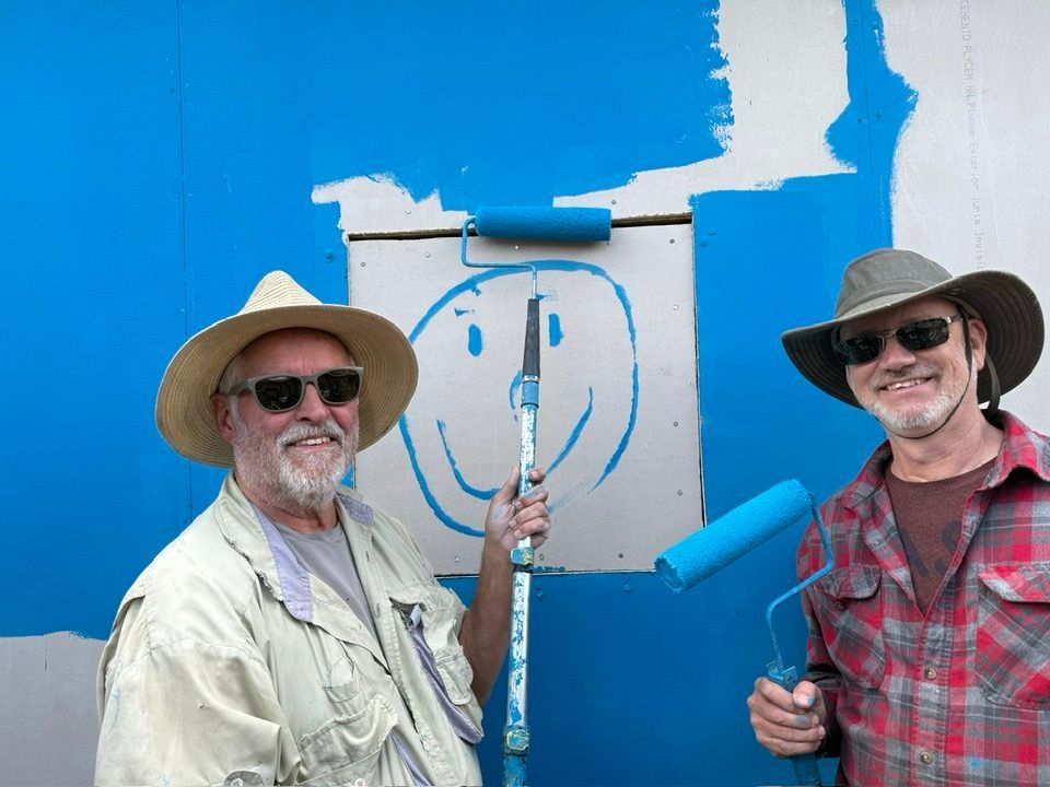 Two people in hats and sunglasses pose smiling in front of a blue wall while holding paint rollers and a smiley face sign.
