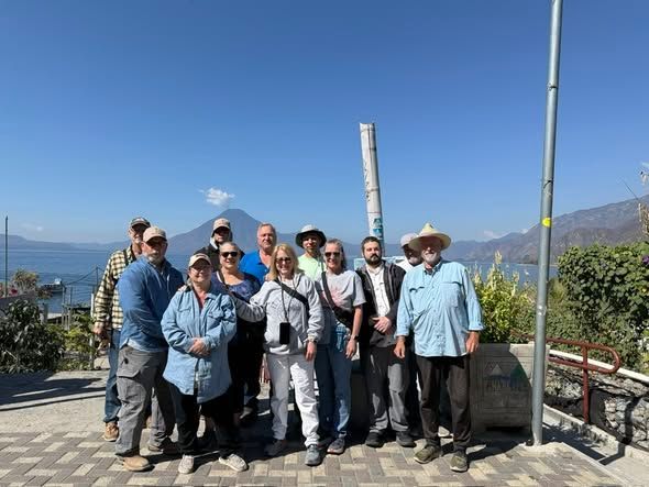 A group of people standing on a paved outdoor terrace overlooking Lake Atitlán and a volcano under a clear blue sky.