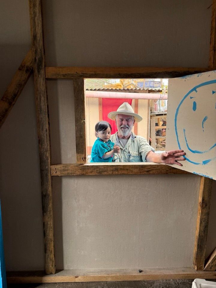 A person holding a child looks through a wooden window frame; a hand holds a sign with a blue smiley face nearby.
