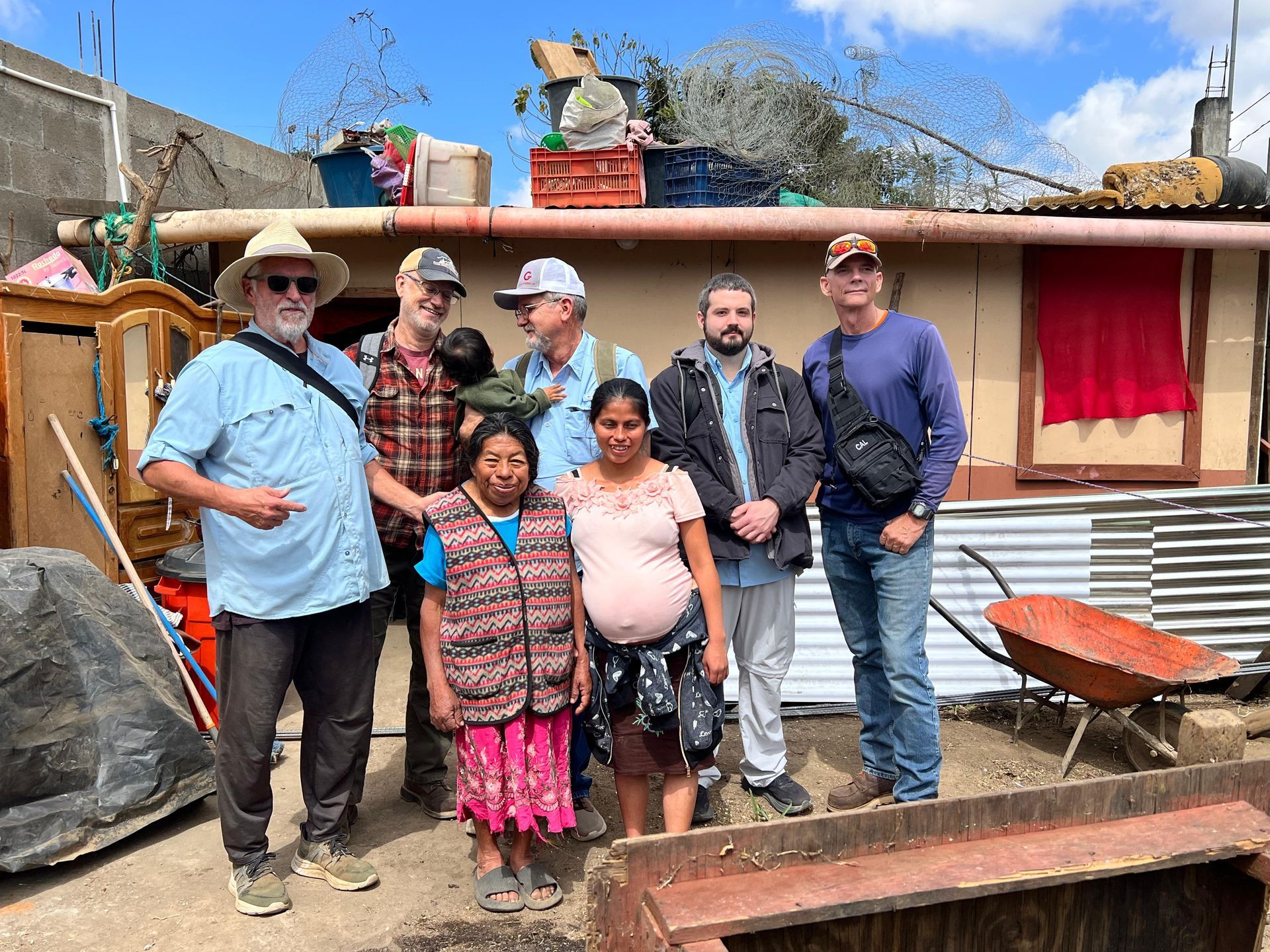 A group of seven people stand smiling in front of a rustic, single-story home with a red cloth hanging on the wall.