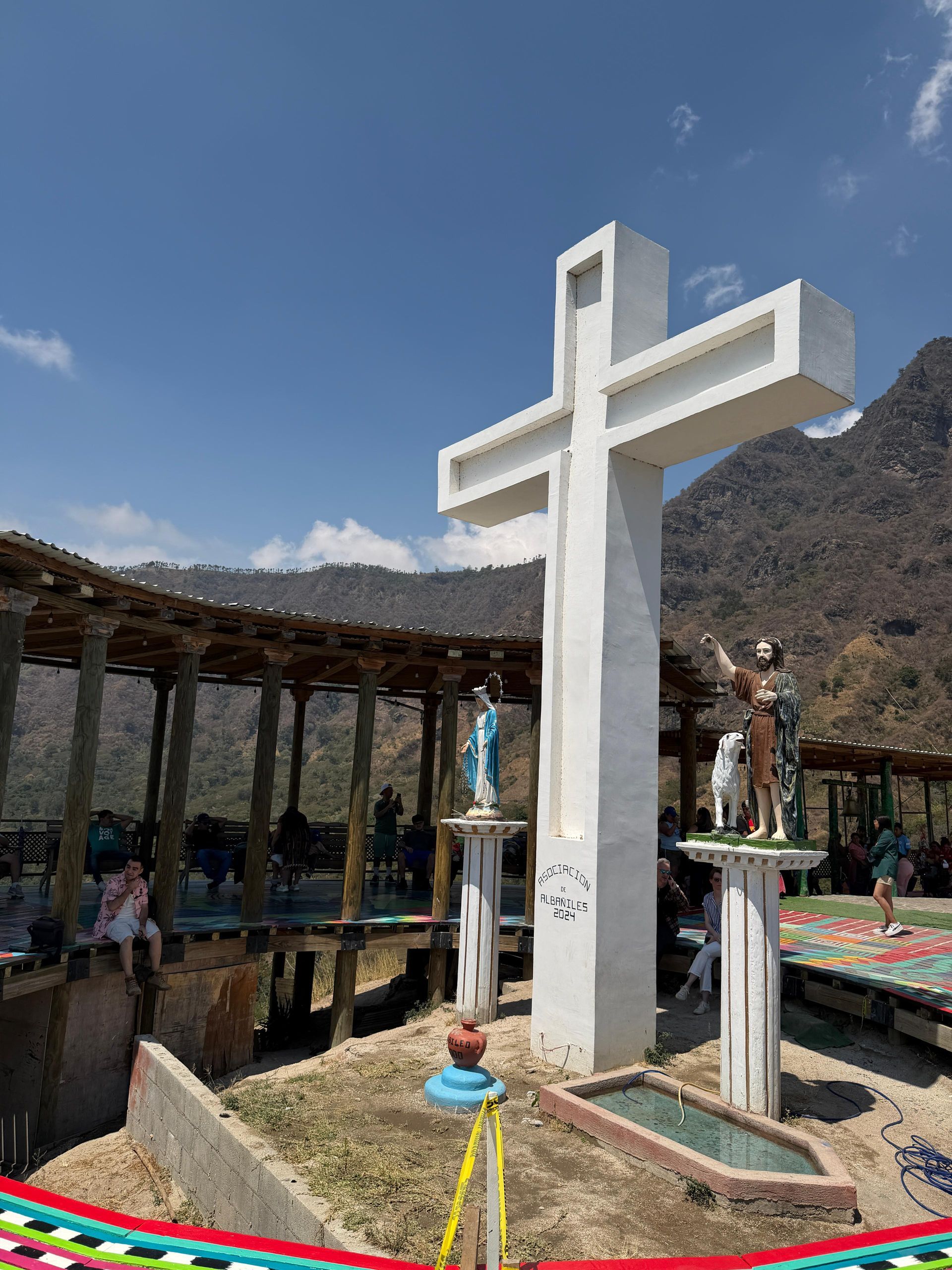 A large white cross stands before a wooden covered walkway, flanked by statues on pedestals against a mountainous backdrop.