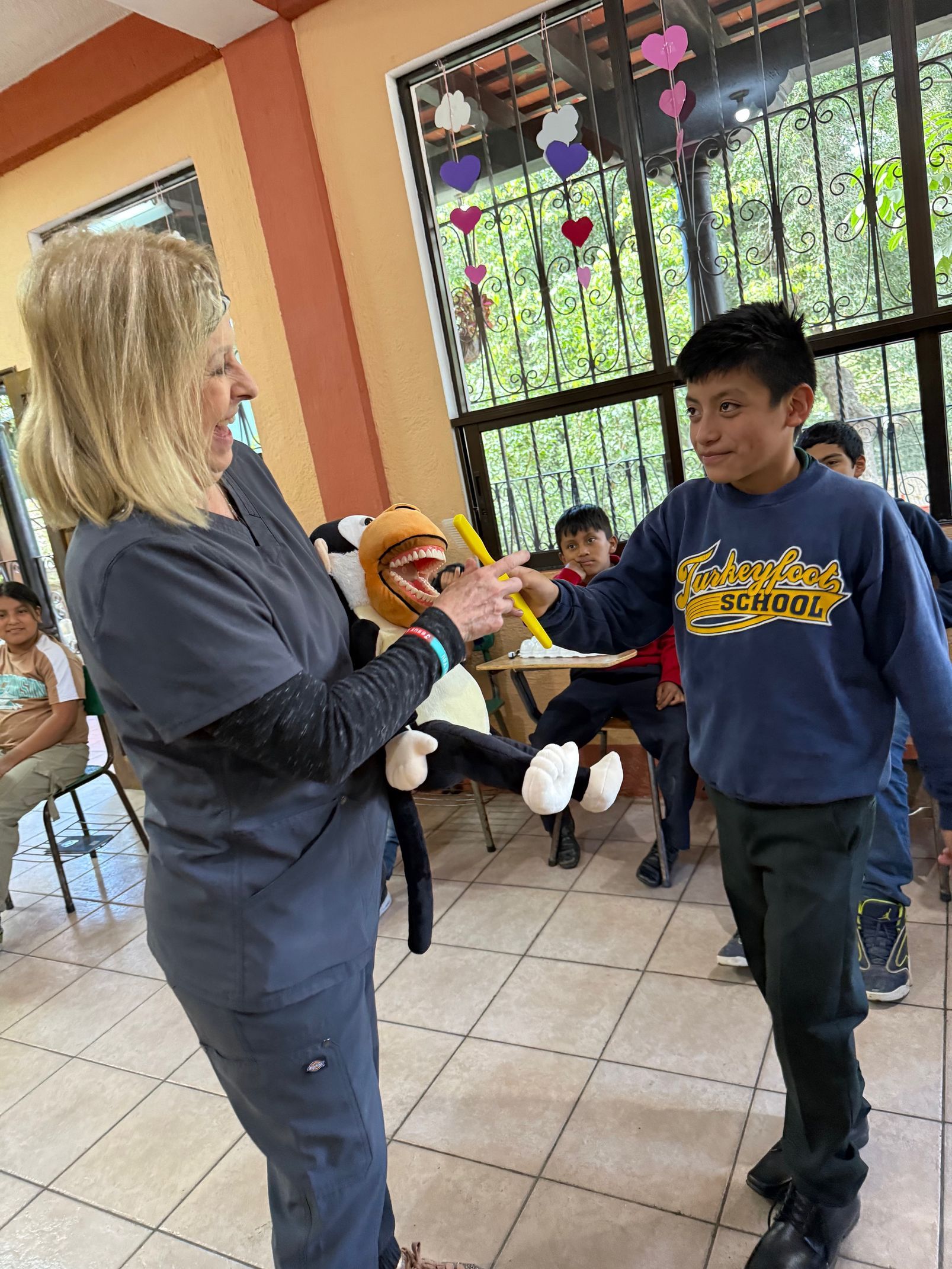 A person in scrubs uses a monkey puppet to teach a student how to brush teeth in a classroom setting.