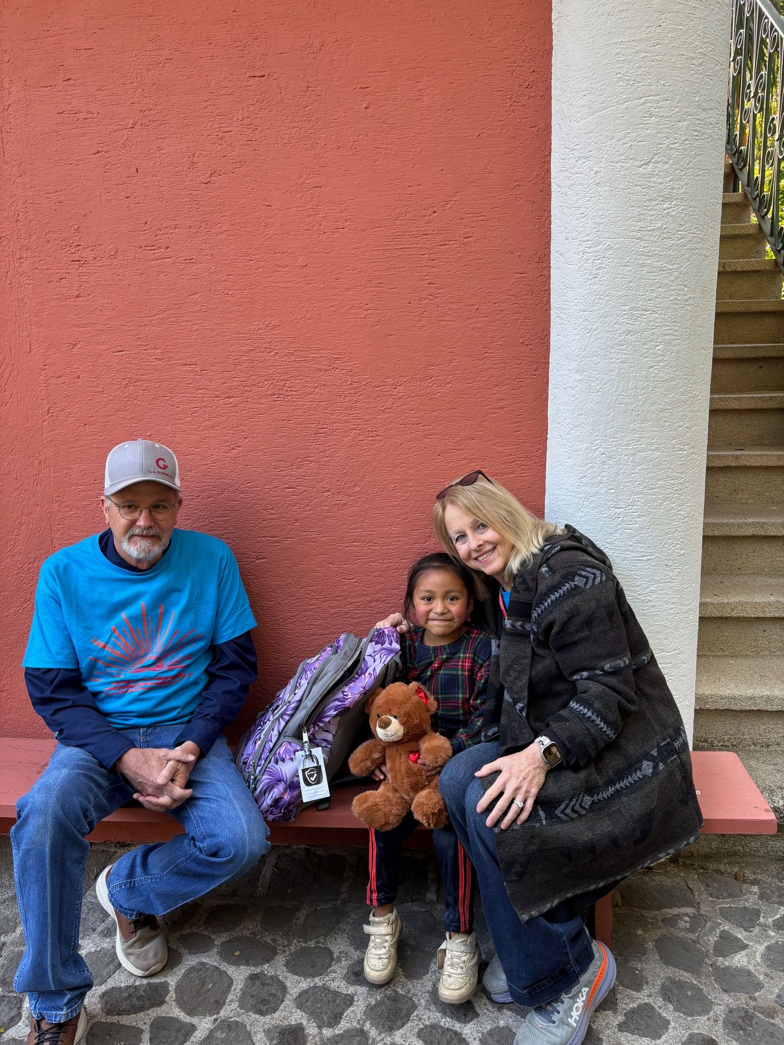 Two adults and a child sitting on a bench against a red wall, with the child holding a teddy bear.