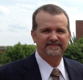 Man in suit and tie outdoors, smiling slightly, with a goatee and short hair.
