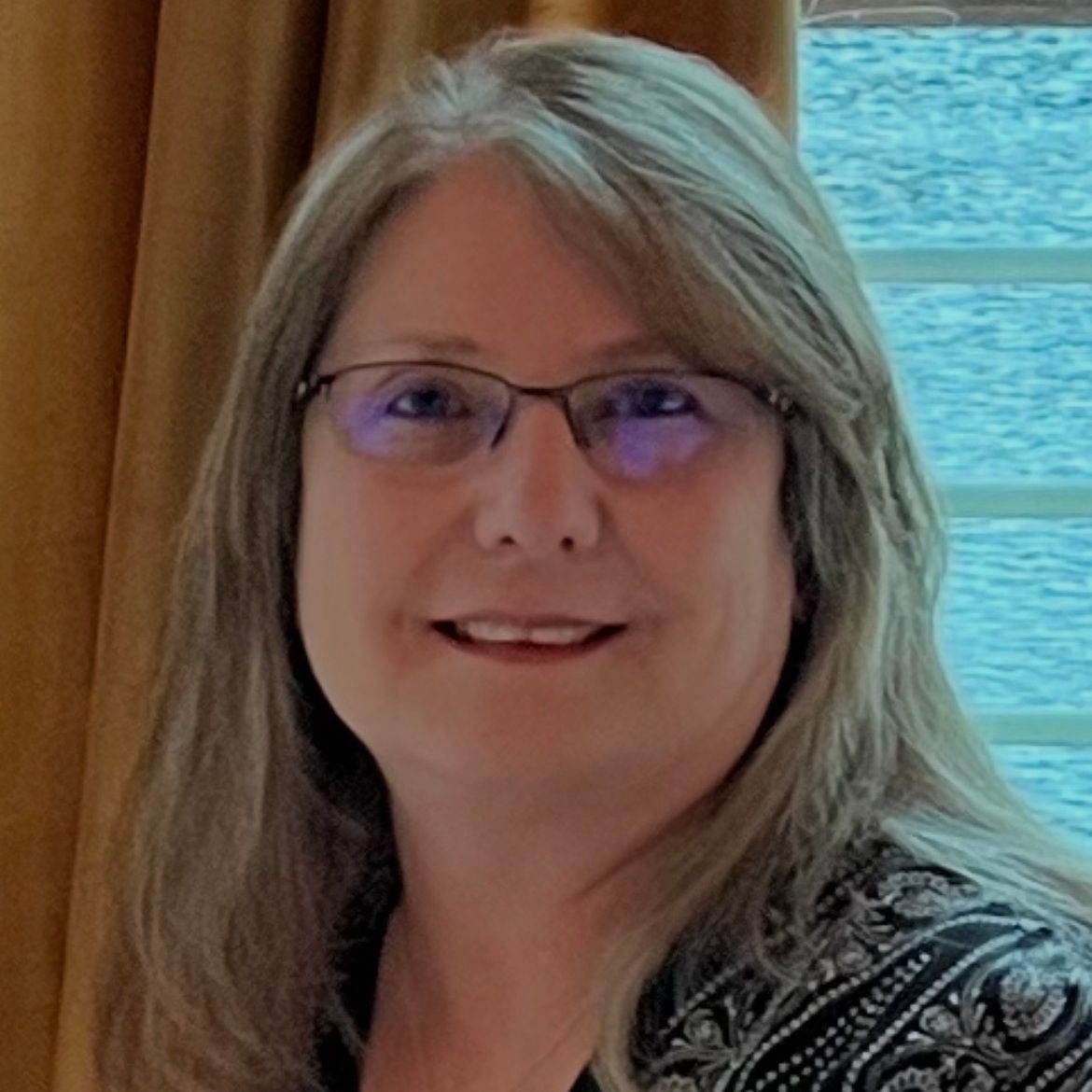 Woman with glasses smiling indoors beside a window with water and curtains in the background