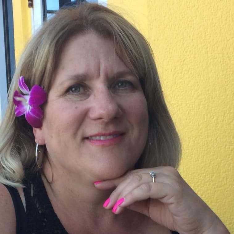 Smiling woman with a purple flower in her hair and pink nails, resting her chin on her hand.