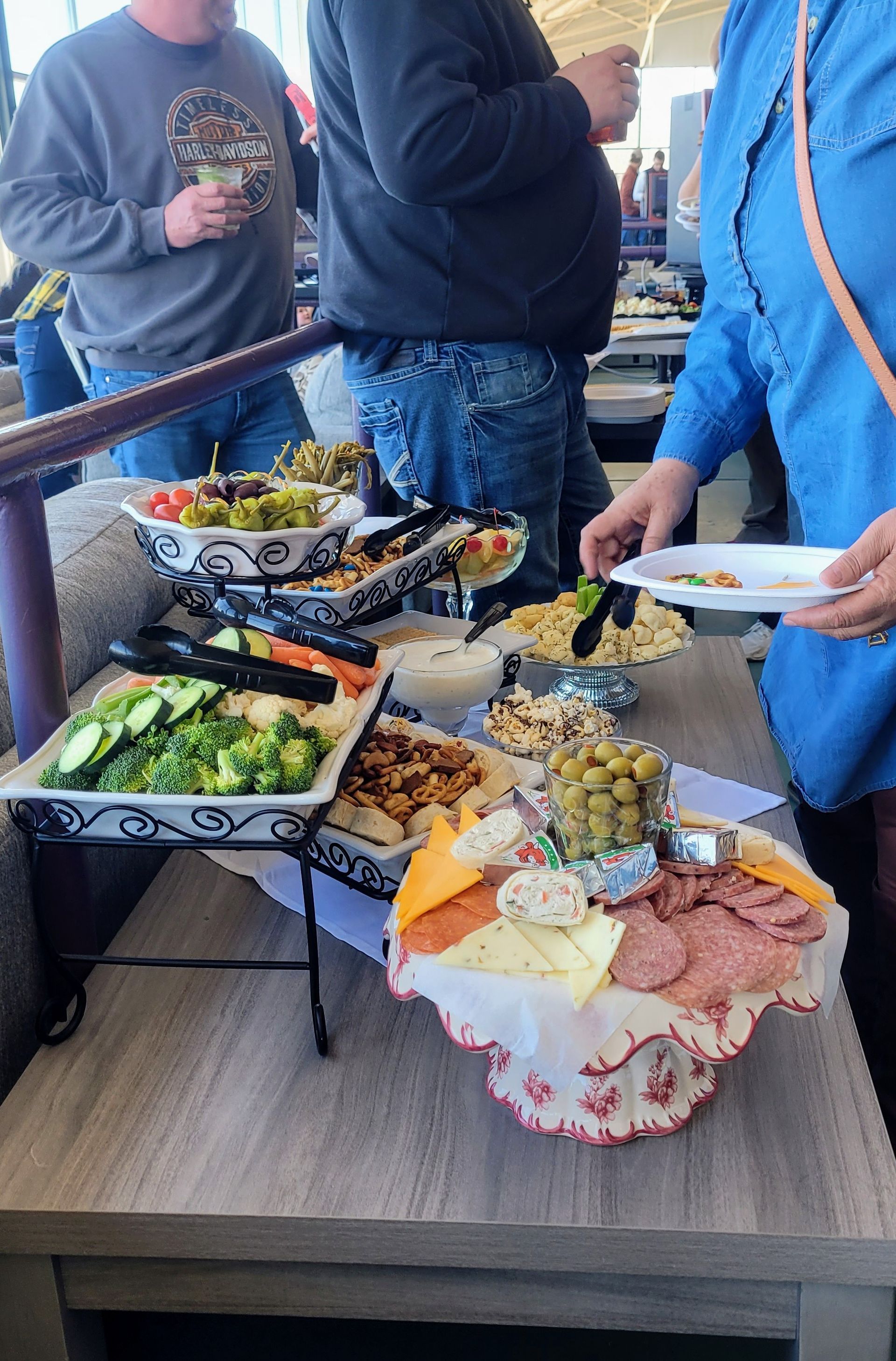 A table with a variety of food on it and people standing around it.