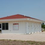A small white house with a red roof is sitting on top of a dirt field.