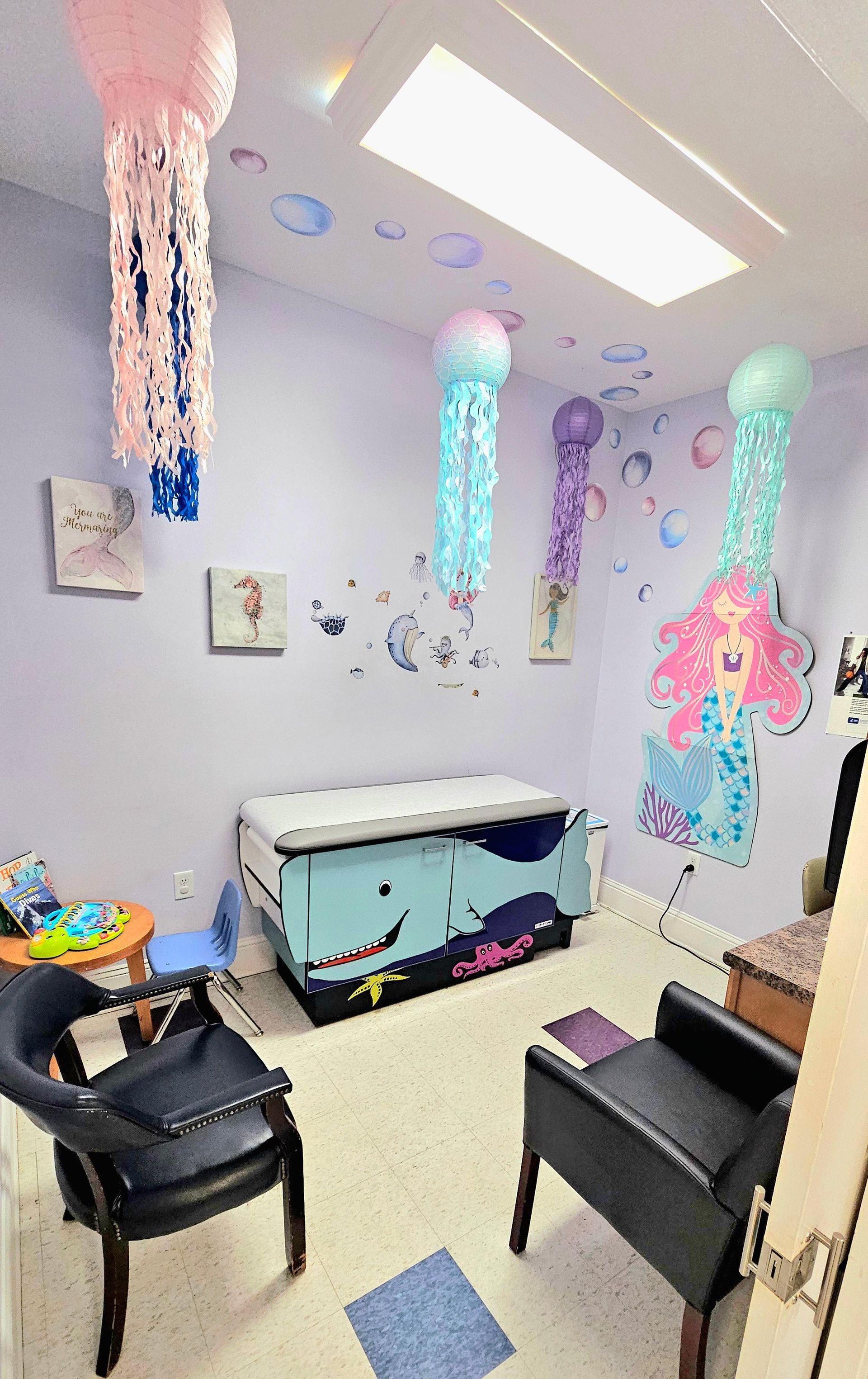 Room decorated with jellyfish decor and mermaid mural, with chairs and examination table.