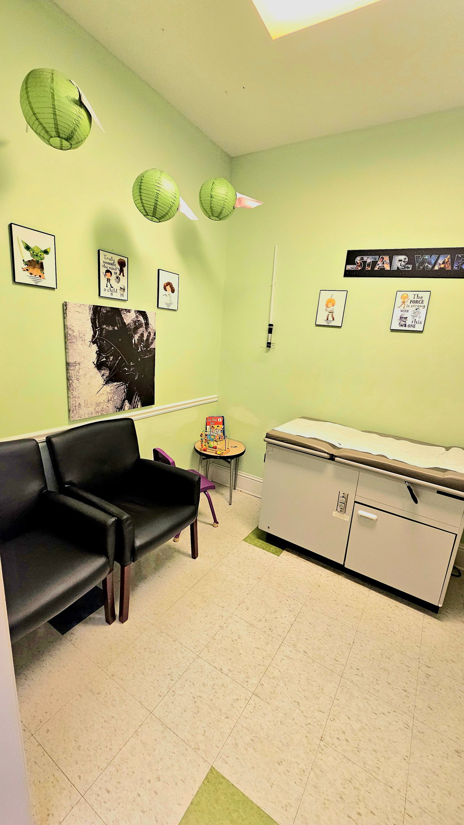 Doctor's examination room with green walls, exam table, waiting chairs, and decorative balloons.